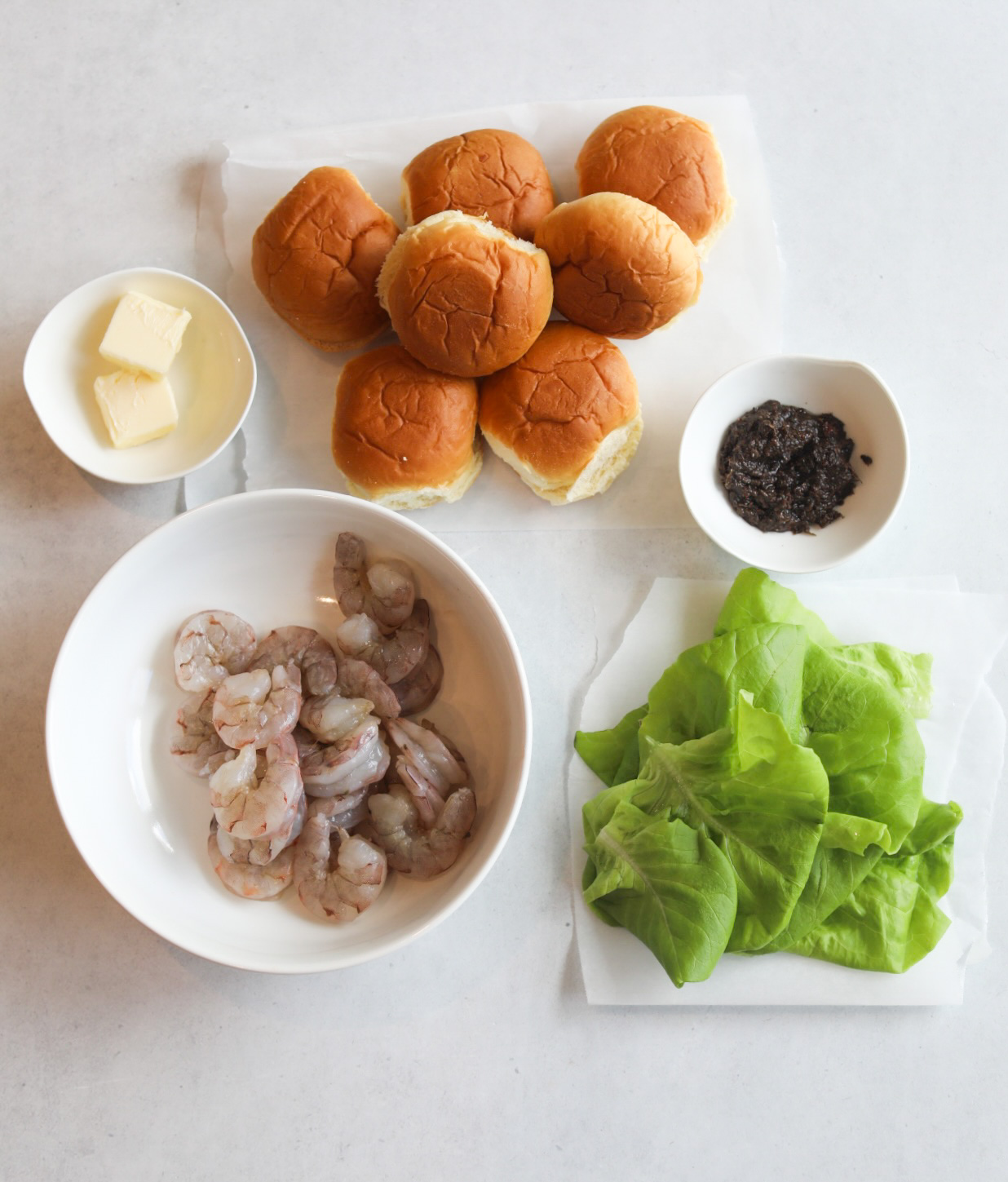 Jerk shrimp slider recipe ingredients. Slider buns are added on white parchment paper, in small white bowls are butter and jerk marinade. Shrimp are in a medium sized bowl with bibb lettuce also on parchment paper.