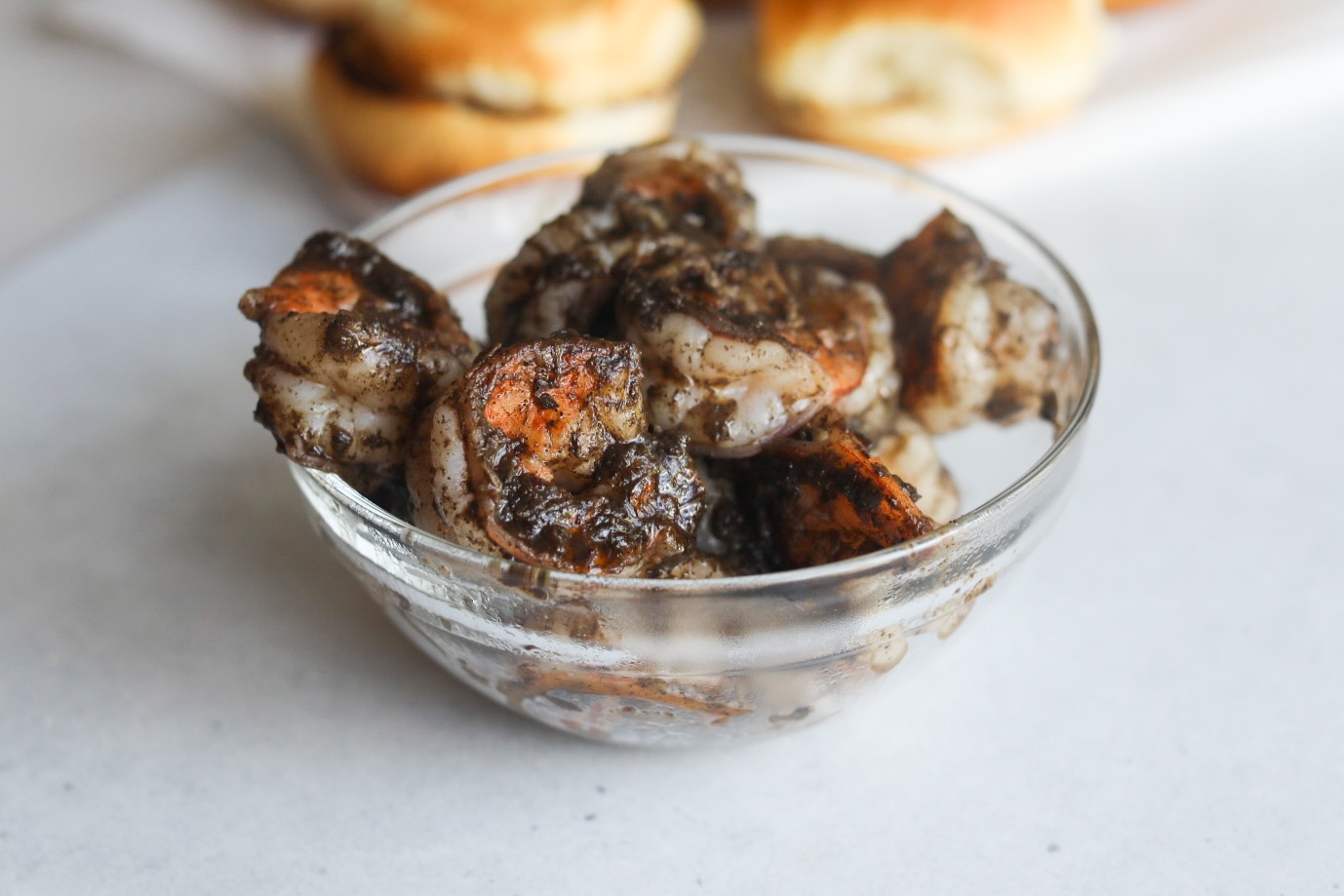 Jerk shrimp cooked with marinade in a small glass bowl cooling off. Slider buns out of focus in the background.