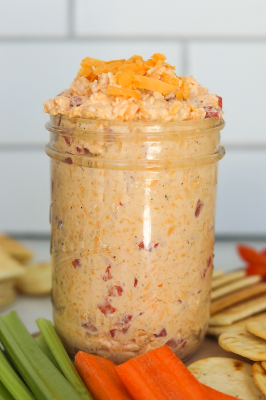 Old Fashioned Pimento Cheese in a small clear glass mason jar with extra shredded cheese added on top. Sliced celery, carrots and crackers added for styling purposes.