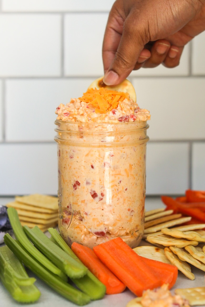 Old fashioned pimento cheese recipe inside of a small mason jar with sliced carrots and celery. Crackers are also added for styling purposes and a hand is scooping out some of the pimento cheese for styling purposes.