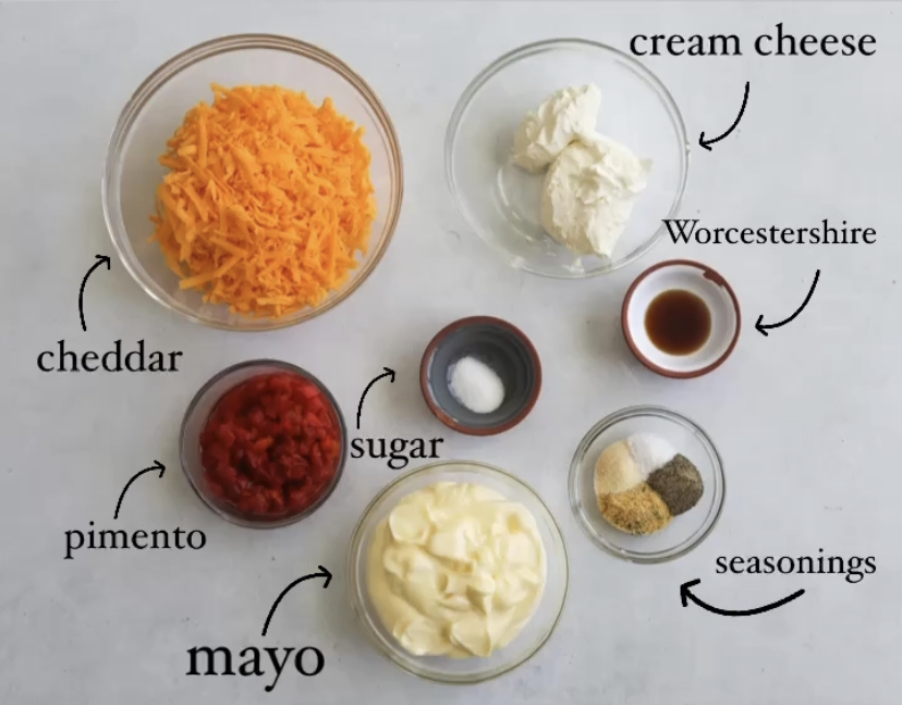 Homemade Pimento Cheese recipe ingredients in a flat lay style. Small bowls are filled with cream cheese, shredded cheddar cheese, pimento, mayo, sugar, seasonings and Worcestershire sauce. Each ingredient is labeled with black text and an arrow.