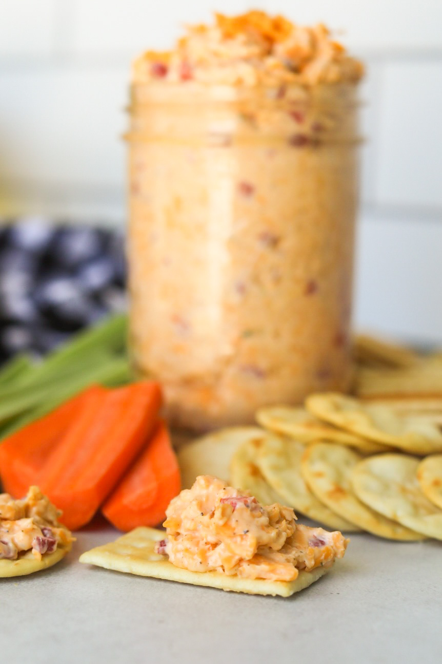 Homemade old fashioned pimento cheese in a mason jar topped with shredded cheddar cheese. Pimento cheese is on a wooden cutting board with sliced carrots and celery as well as assorted crackers added for styling purposes. Two crackers are in focus in front of the staged scene with scooped out pimento cheese on top of each cracker.