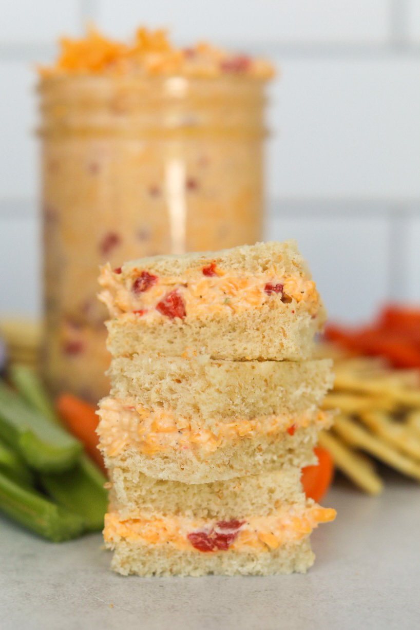 Old fashioned pimento cheese recipe made into small square sandwiches with bread. In the background out of focus are sliced celery and carrot sticks as well as crackers.