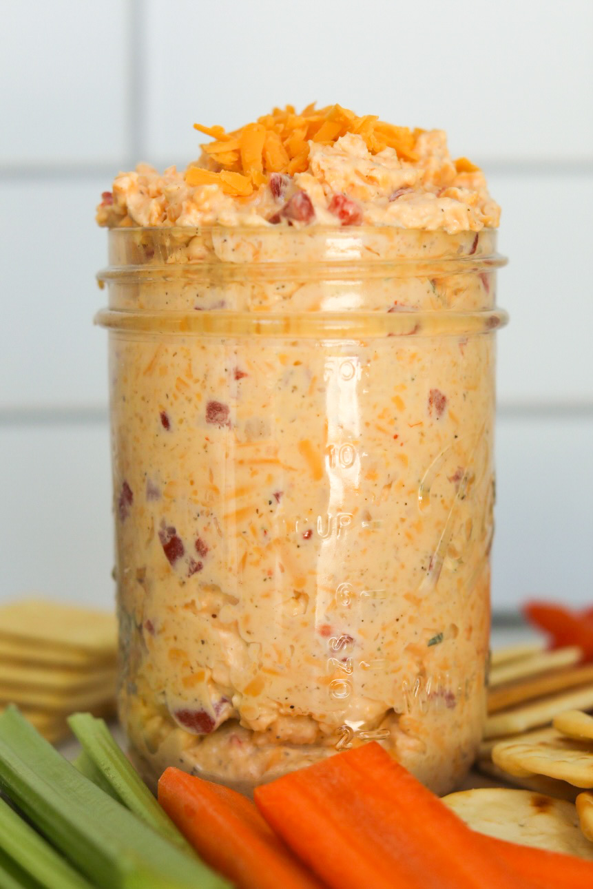 Homemade old fashioned pimento cheese in a mason jar topped with shredded cheddar cheese. Pimento cheese is on a wooden cutting board with sliced carrots and celery as well as assorted crackers added for styling purposes. Two crackers are in focus in front of the staged scene with scooped out pimento cheese on top of each cracker.
