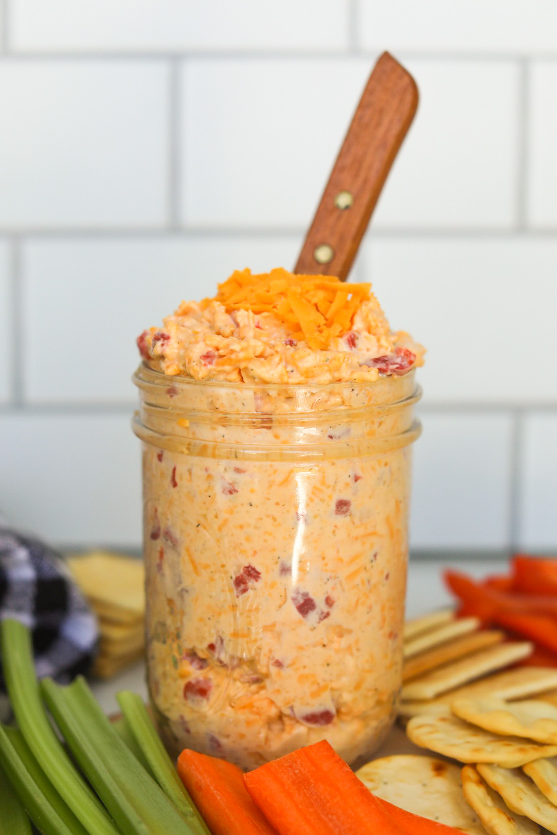 Old Fashioned Pimento Cheese in a small clear glass mason jar with extra shredded cheese added on top. Sliced celery, carrots and crackers and a brown knife added for styling purposes.