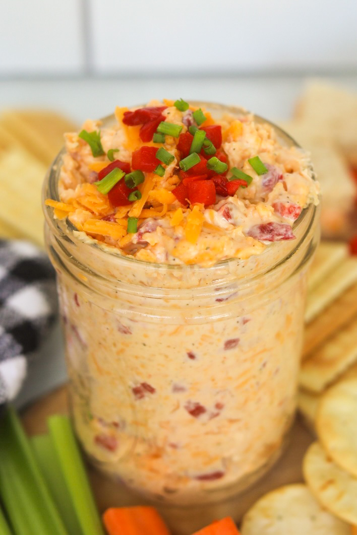 Homemade pimento cheese topped with shredded cheddar cheese, pimento and green onions. Sliced celery and carrots as well as a black and white kitchen towel added for styling purposes.