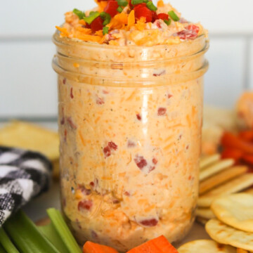 Pimento cheese filled in a glass mason jar with shredded cheddar, pimento and green onions topped for styling purposes. Silver spoon added to scoop pimento cheese with crackers, sliced celery and carrots and a black and white checkered towel added for styling purposes.