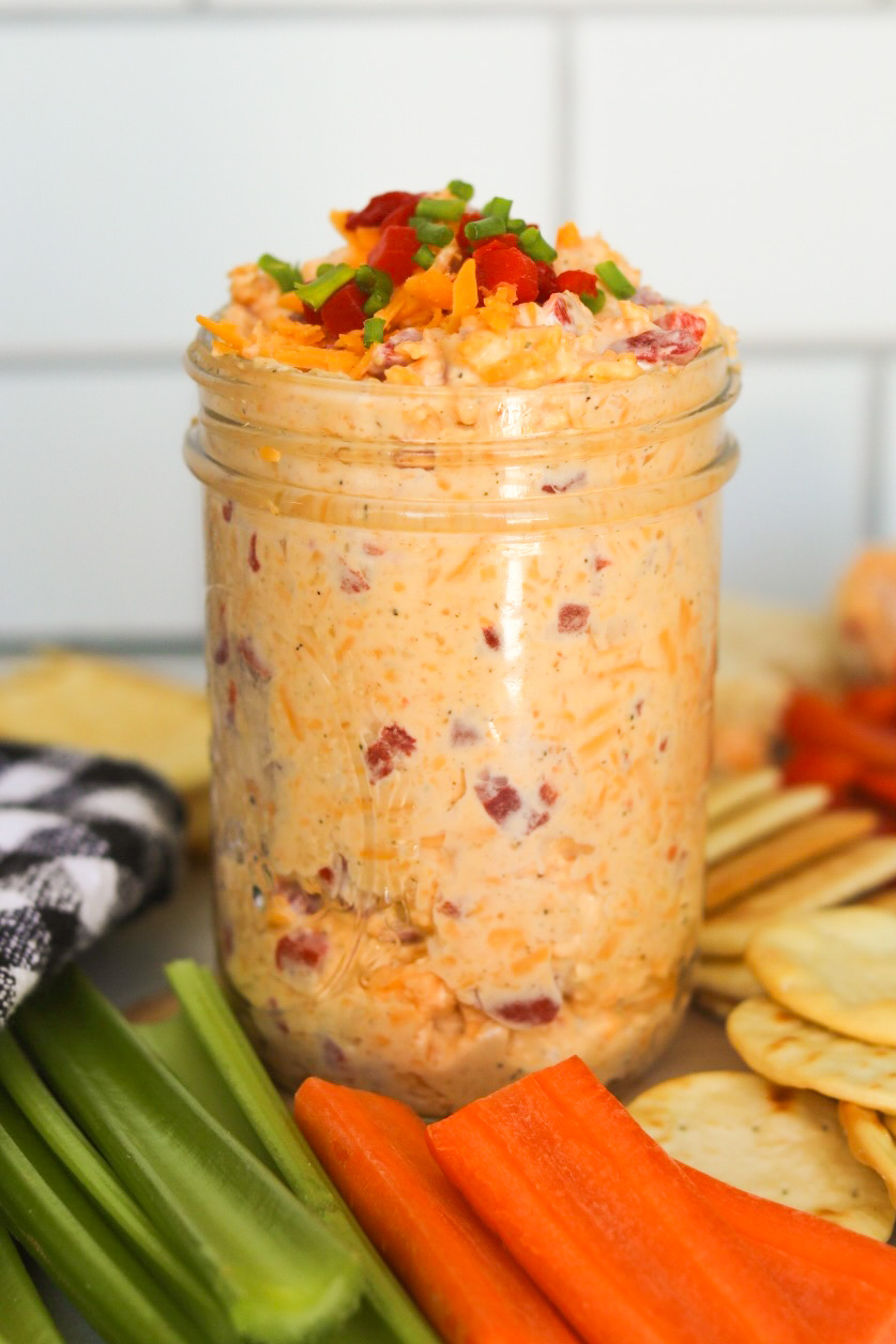 Pimento cheese filled in a glass mason jar with shredded cheddar, pimento and green onions topped for styling purposes. Silver spoon added to scoop pimento cheese with crackers, sliced celery and carrots and a black and white checkered towel added for styling purposes.