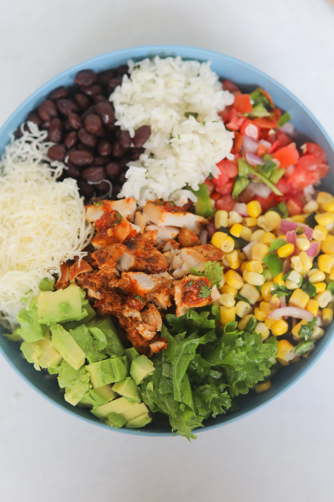 Burrito Bowl in blue bowl with corn salsa, pico de gallo, white rice, black beans, Monterey jack cheese, avocado, lettuce and pollo asado chicken.