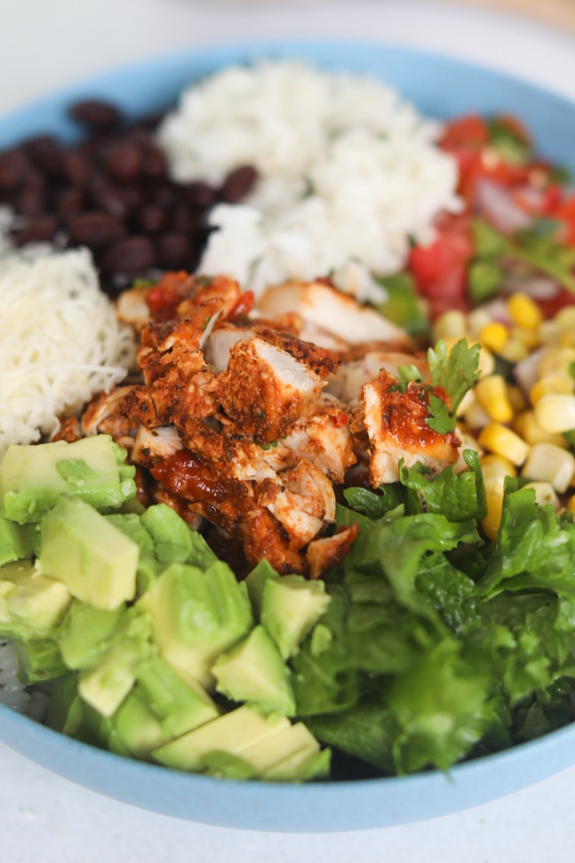 Bowl of deconstructed burrito bowl. In focus is chopped avocado, green lettuce, corn salsa, cheese and pollo asado chicken.