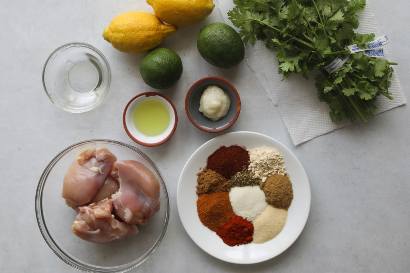 Pollo asado marinade ingredients. From left to right, bunch of cilantro, lemons, limes, small bowls of garlic paste, oil, vinegar, a glass bowl of chicken thighs and a small white plate with seasonings to make marinade in a flat lay.