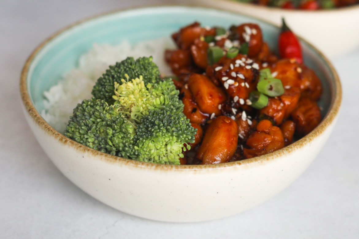 Spicy Chicken Teriyaki plated in a blue bowl with broccoli and white rice. Plated dish is topped with chopped green onion, sesame seeds and a red chili pepper added for styling purposes.