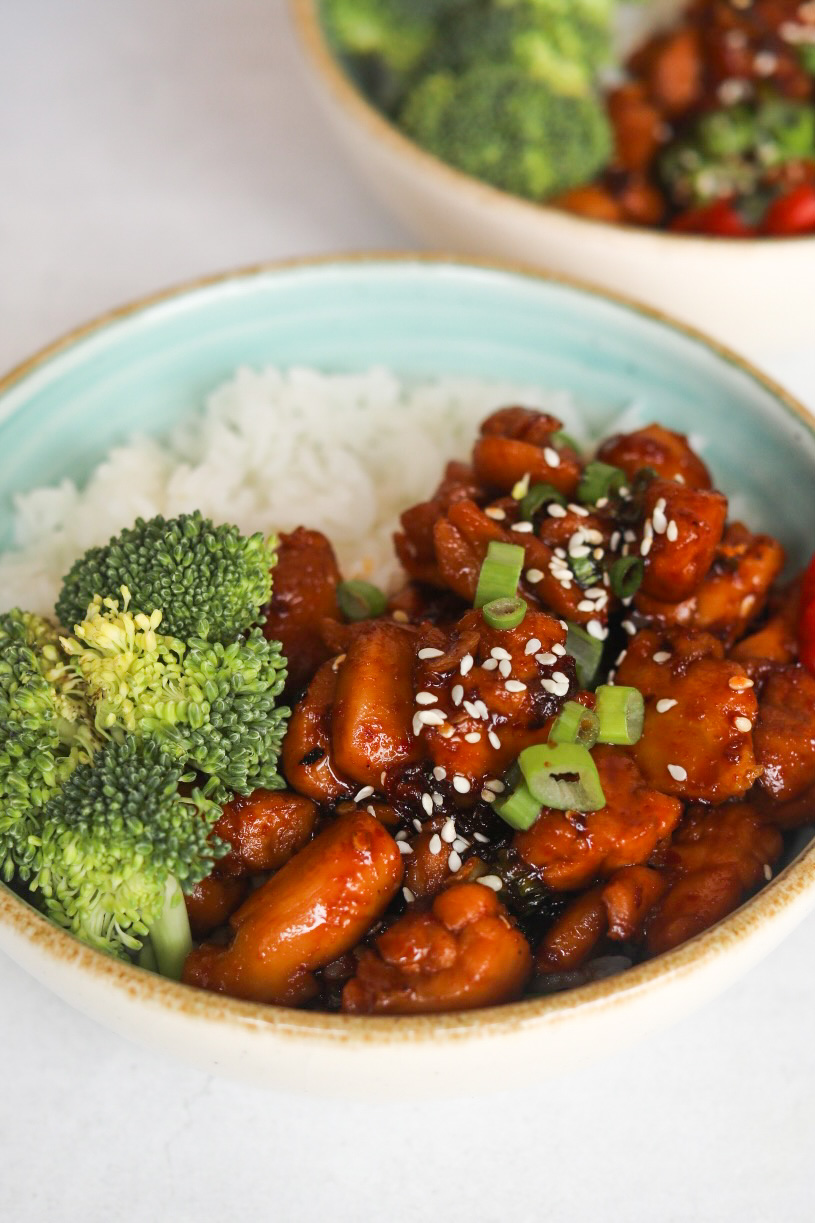 Spicy Chicken Teriyaki plated in a blue bowl with broccoli and white rice. Plated dish is topped with chopped green onion, sesame seeds.