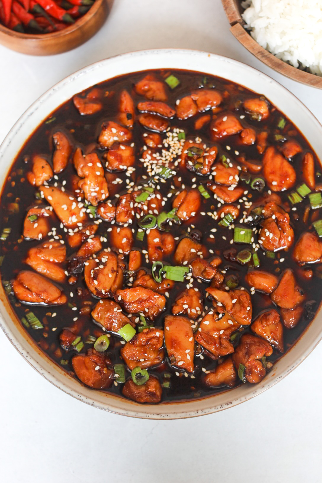 Spicy Chicken Teriyaki in a large serving bowl topped with sesame seeds and green onions. White rice in a wooden bowl and red chili peppers in a small wooden bowl is added for styling purposes in the top left and right corners of the image.