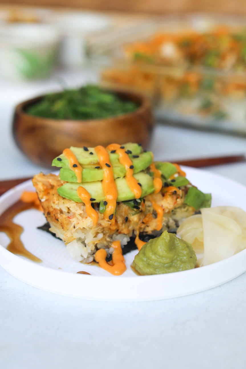 Sushi bake with spicy crab plated on a small white plate topped with avocado, spicy mayo and black sesame seeds. Out of focus in a wooden bowl is seaweed salad and casserole dish with sushi bake.