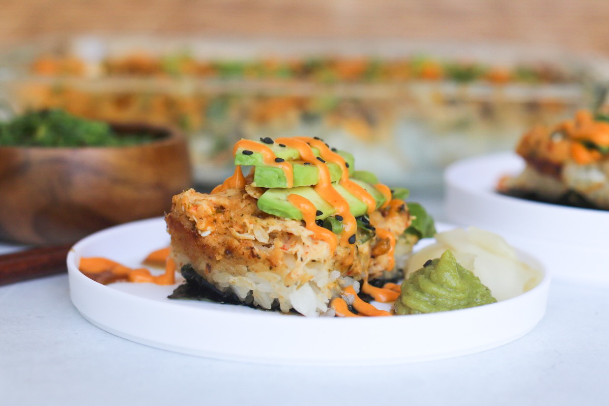 Plated sushi bake on a white plate drizzled with spicy mayo, avocado and black sesame seeds.