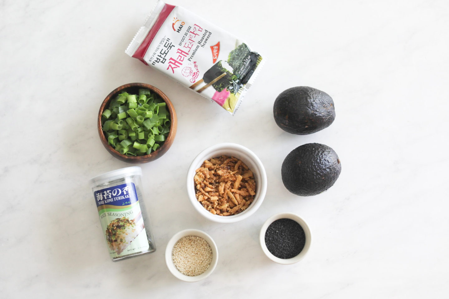 Additional toppings for sushi bake with additional toppings. From left to right in a flat lay style, seaweed sheets packaged, wooden bowl of green onions, bottled furikake, small white bowl with sesame seeds, small white bowl with fried green onions, small white bowl with black sesame seeds and two avocados.