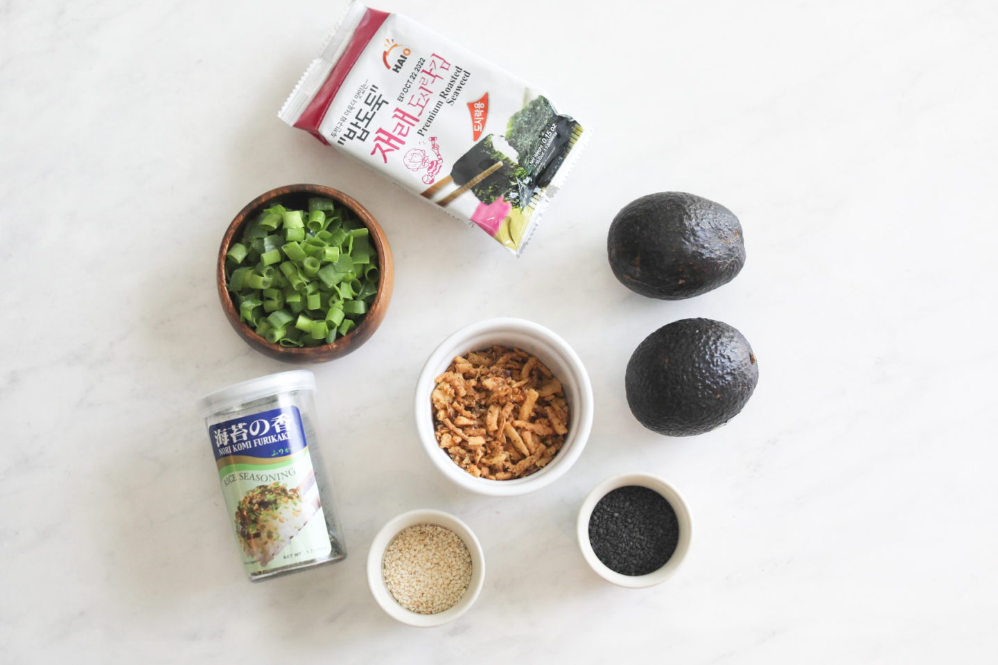 spicy crab sushi bake Additional toppings for sushi bake with additional toppings. From left to right in a flat lay style, seaweed sheets packaged, wooden bowl of green onions, bottled furikake, small white bowl with sesame seeds, small white bowl with fried green onions, small white bowl with black sesame seeds and two avocados.