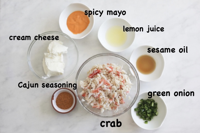 Sushi bake ingredients on a flay late in small white and clear bowls. From left to right, small bowl of spicy mayo, cream cheese, Cajun seasoning, crab meat, jalapenos, sesame oil and lemon juice.