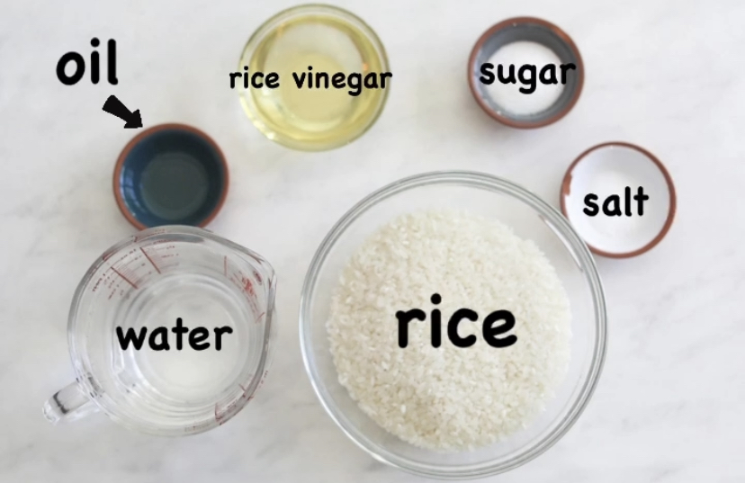Sushi rice ingredients for sushi bake. Small bowls from left to right of rice vinegar, oil, water, rice, salt and sugar.