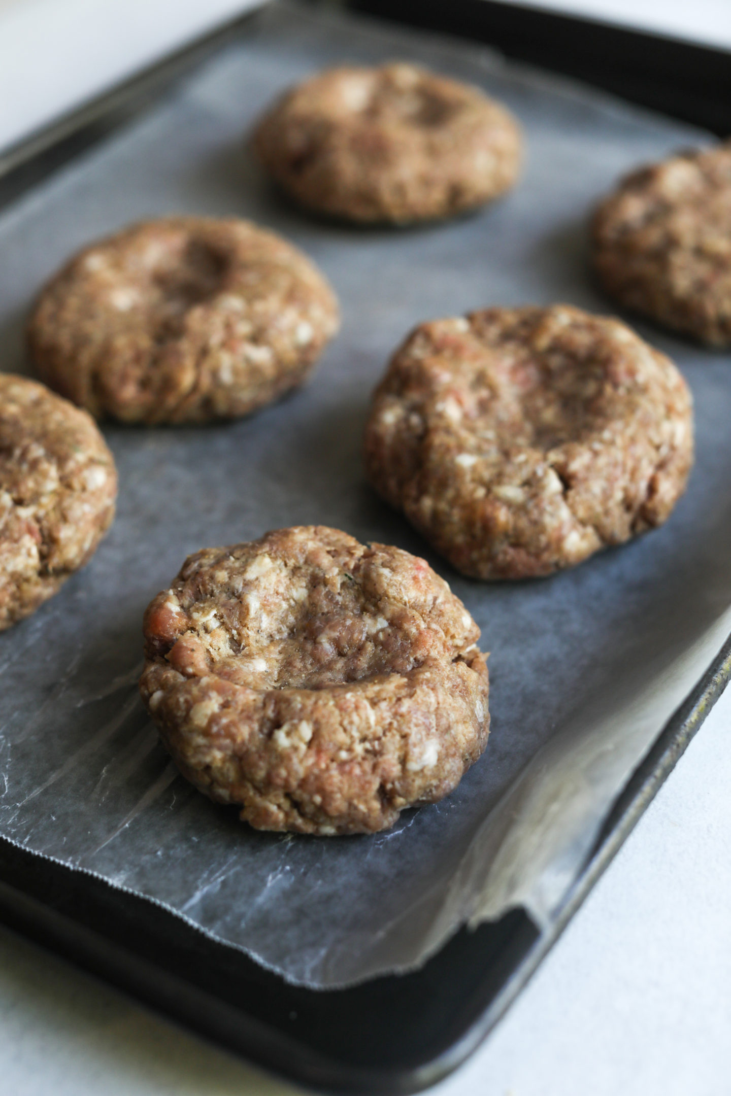 Six raw burger patties with seasoning on a baking sheet lined with wax paper.