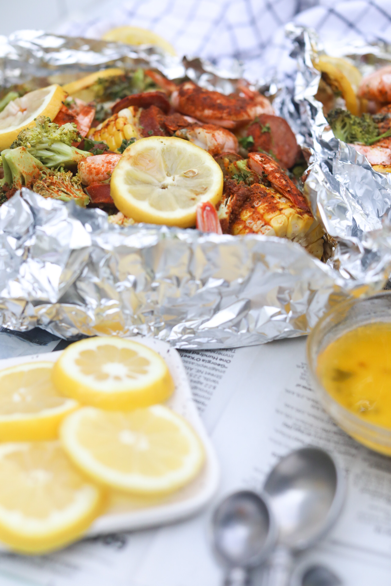 Baked shrimp boil complete sat under newspaper for styling and eating purposes. Small white plate with sliced lemons and a glass bowl filled with garlic butter sauce and measuring spoons, black and white kitchen towel added for styling purposes.