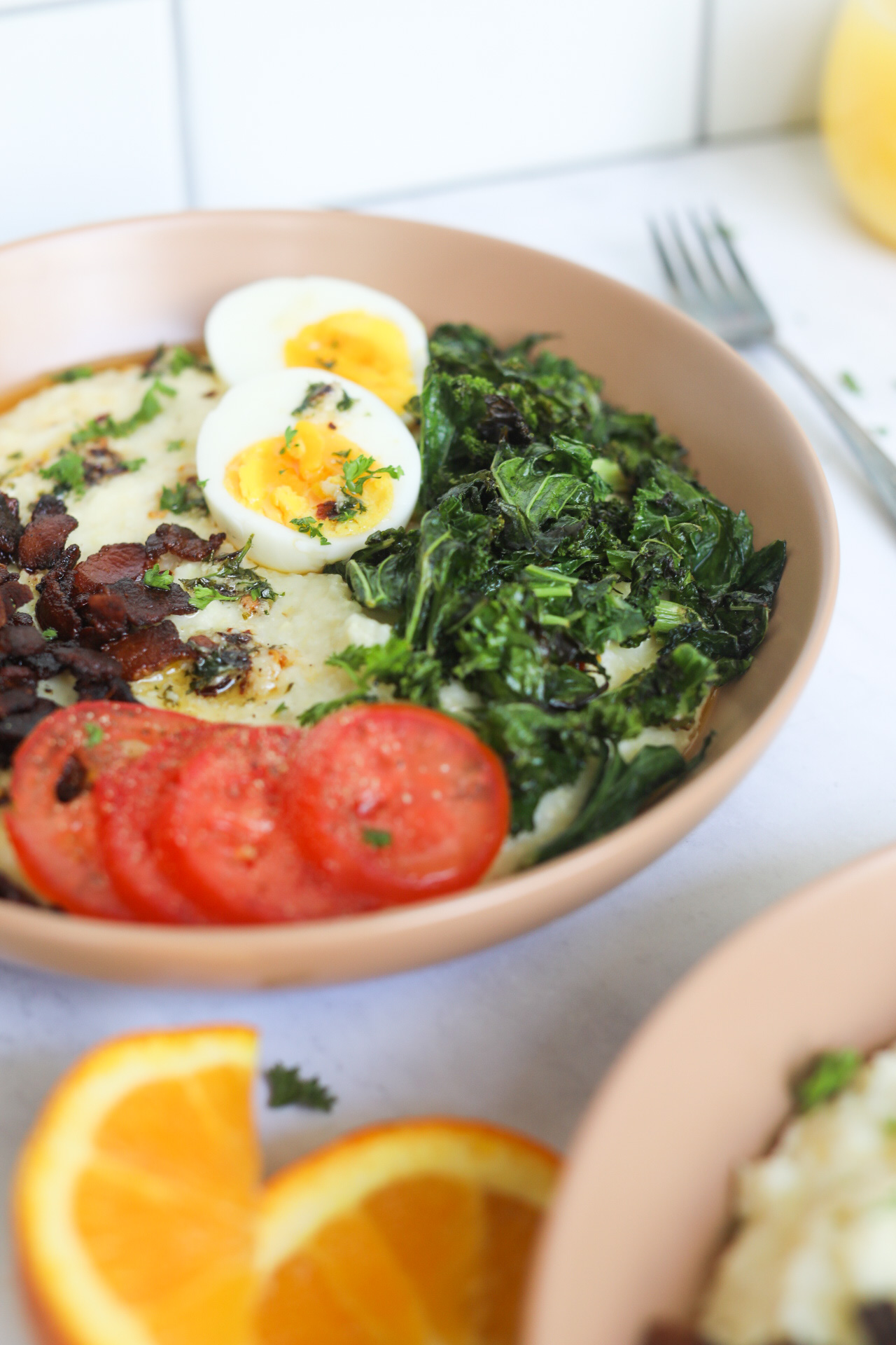 Southern Grits and Greens Bowl, finished recipe in a light tan bowl. Sliced oranges, fork and wine glass filled with orange juice for styling purposes.