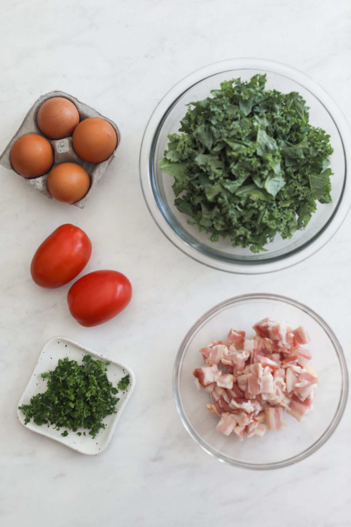 Southern Grits and Greens ingredients of additional items. Flat lay of four brown eggs in a carton, two roma tomatoes, small white plate of chopped parsley, glass bowl of chopped bacon and glass bowl of kale.