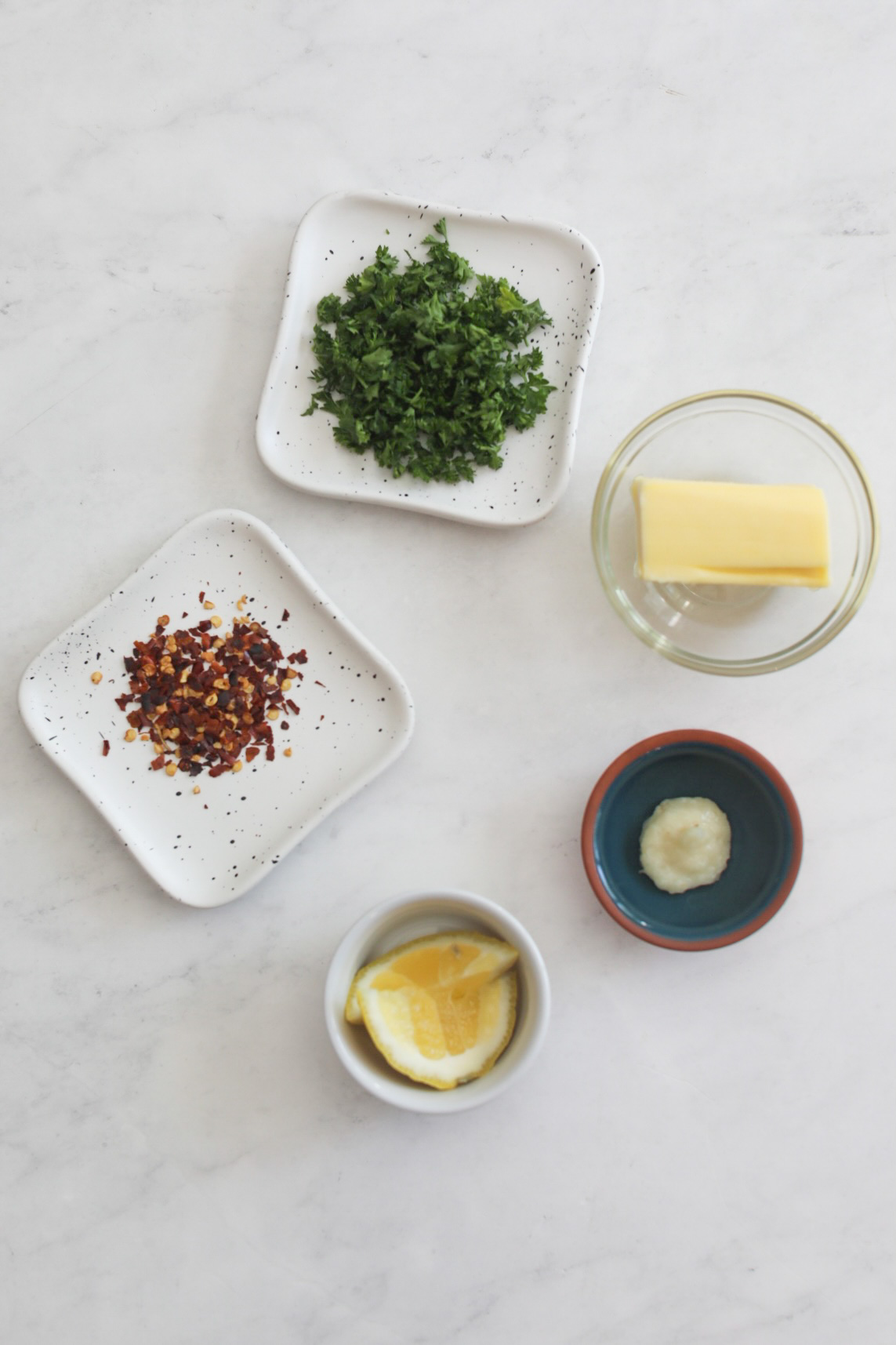 Garlic butter sauce ingredients in a flat lay style. Two white plates with black spots are filled with chopped parsley and crushed red pepper flakes. A small glass bowl of butter, two lemon slices and garlic paste are added as well.