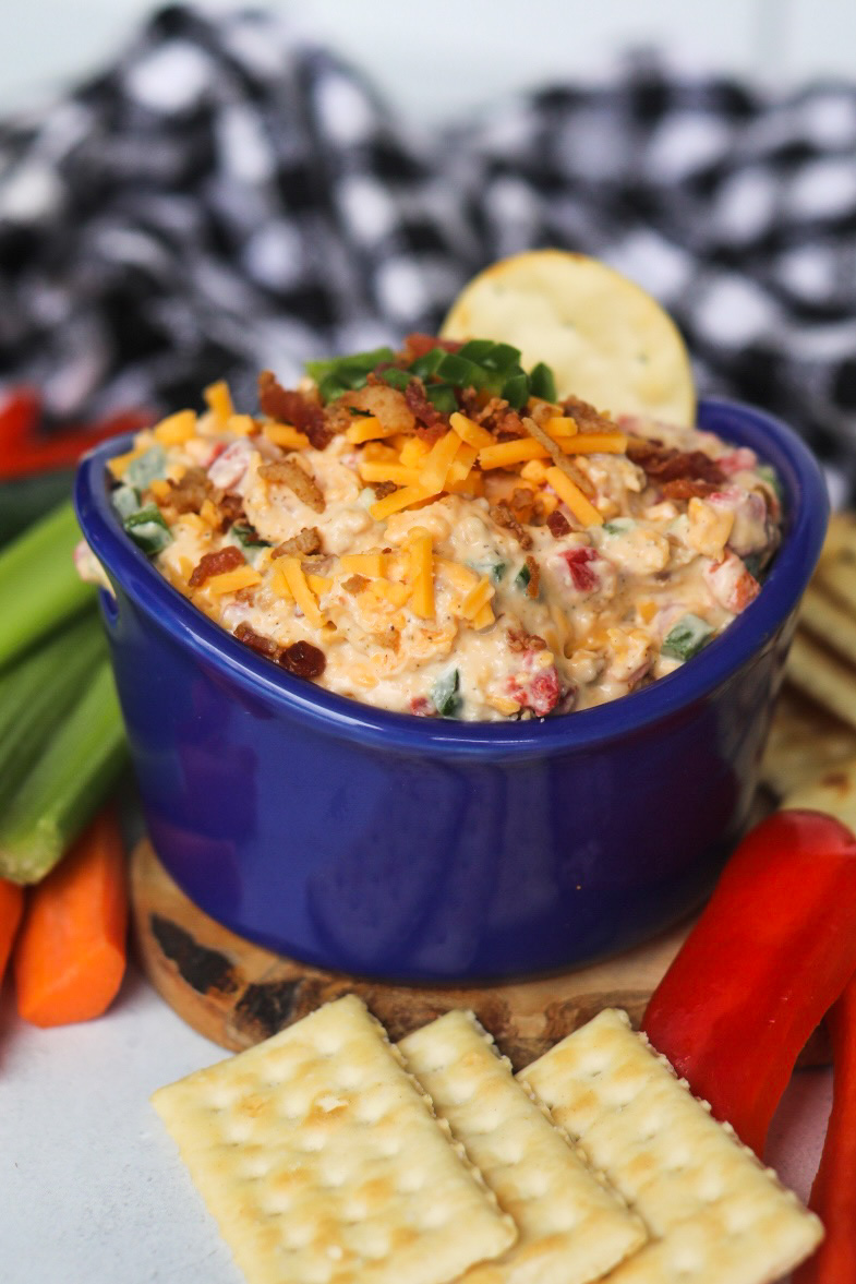 Spicy pimento cheese in a blue bowl topped with shredded cheddar, crumbled bacon bits and diced jalapeno. Crackers, sliced carrots, celery and red bell peppers and black and white kitchen towel added for styling purposes.