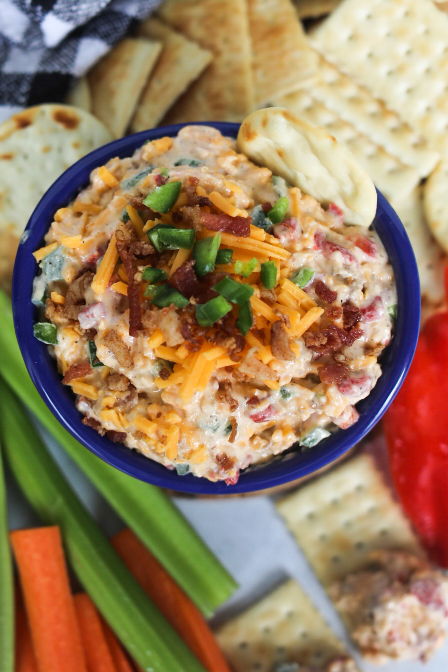 Jalapeno pimento cheese finished in a blue bowl surrounded by crackers, celery and carrot sticks.