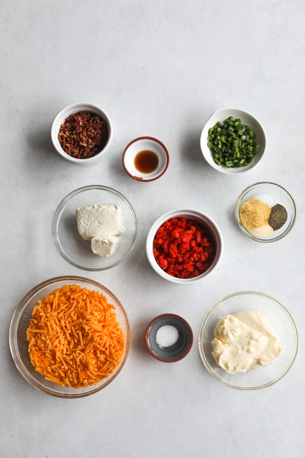 Jalapeno pimento cheese recipe ingredients in a flat lay. Small clear and white bowls are filled with crumbled bacon bits, diced jalapeno, Worcestershire sauce, cream cheese, shredded cheddar cheese, sugar, seasoning blend, pimento and mayonnaise.