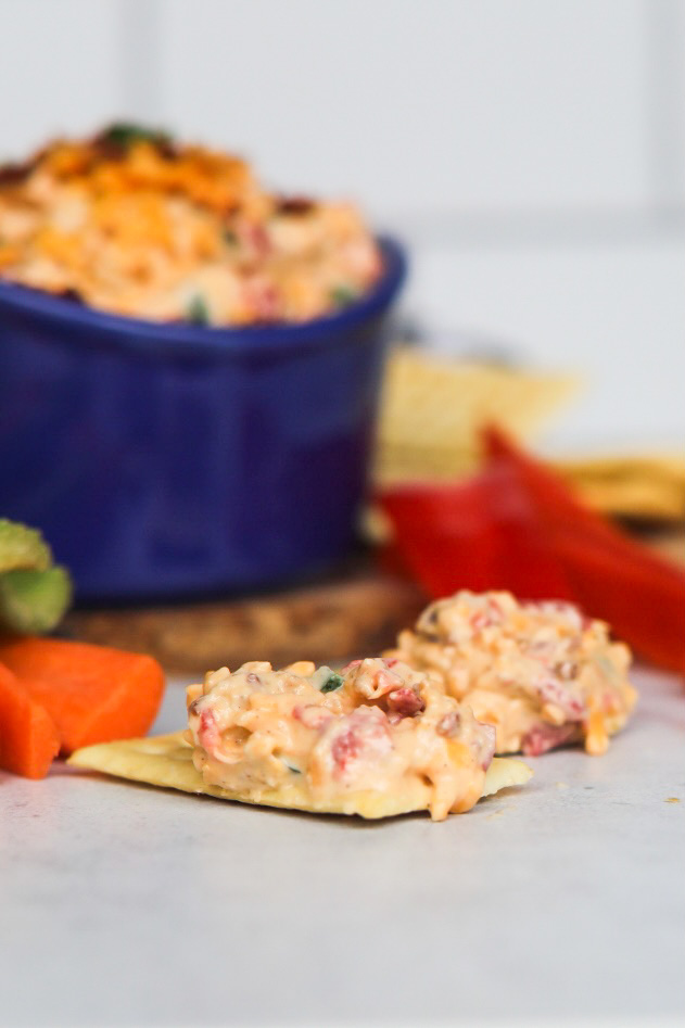 Crackers lying flat with pimento cheese spreaded on top. Our of focus is a blue bowl with jalapeno pimento cheese. Sliced carrots, celery and red bell peppers added for styling purposes.