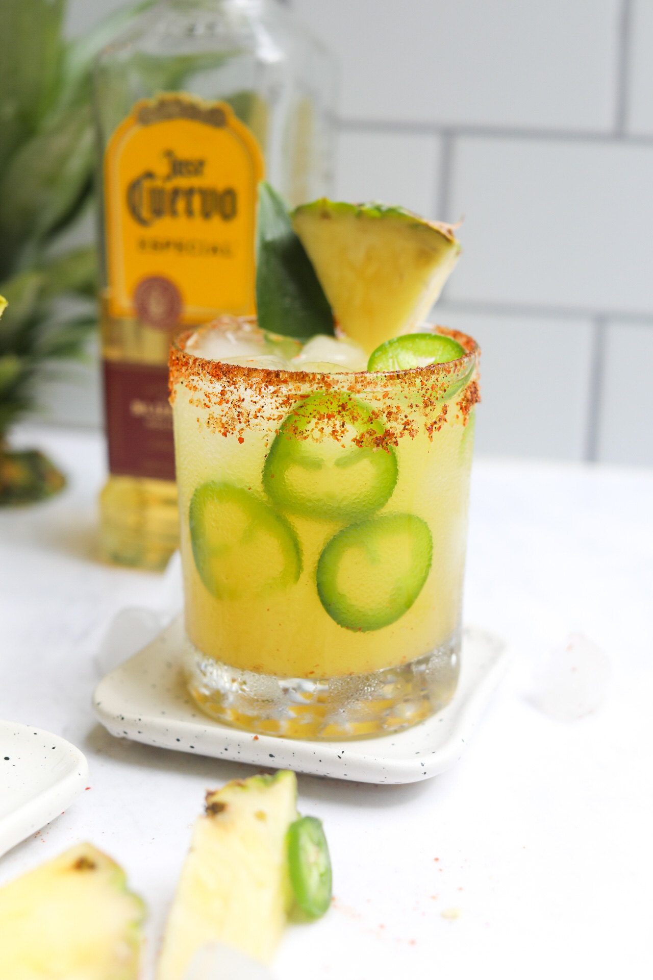 Margarita in a short glass dressed with sliced jalapenos, pineapple slice and pineapple leaf. Tequila bottle is behind the cocktail as well as the top of a fresh pineapple for styling purposes.