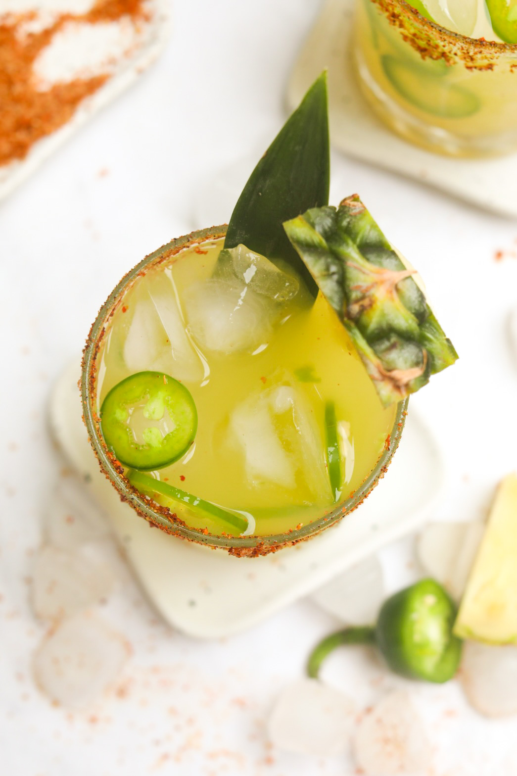 Jalapeno Pineapple Margarita in a short glass, top- down angle, dressed with sliced jalapenos, pineapple slice and pineapple leaf. Tequila bottle is behind the cocktail as well as the top of a fresh pineapple for styling purposes.