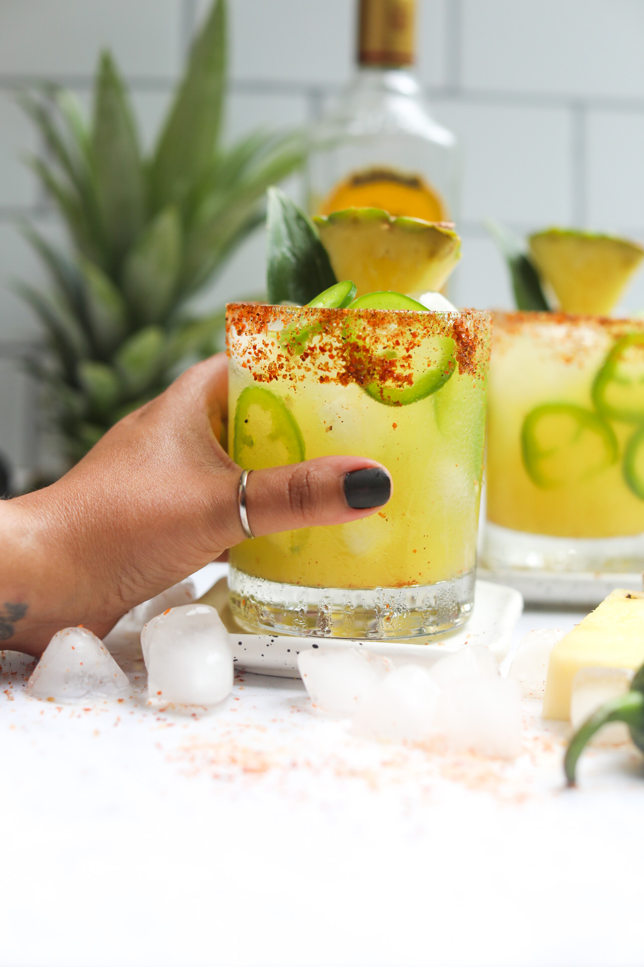 Margarita in a short glass dressed with sliced jalapenos, pineapple slice and pineapple leaf. Tequila bottle is behind the cocktail as well as the top of a fresh pineapple for styling purposes. A left hand is holding the cocktail glass with thumb showing black fingernail polish and a gold ring around the thumb.