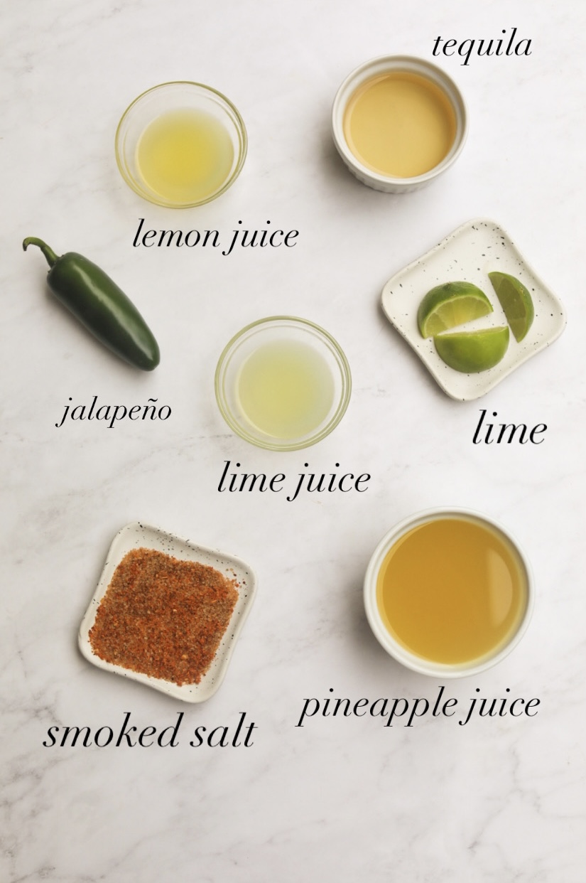 Recipe ingredients on a flat lay. Small bowls are filled and labeled. Tequila, lemon juice, jalapeno, lime juice, sliced limes, smoked salt and pineapple juice.