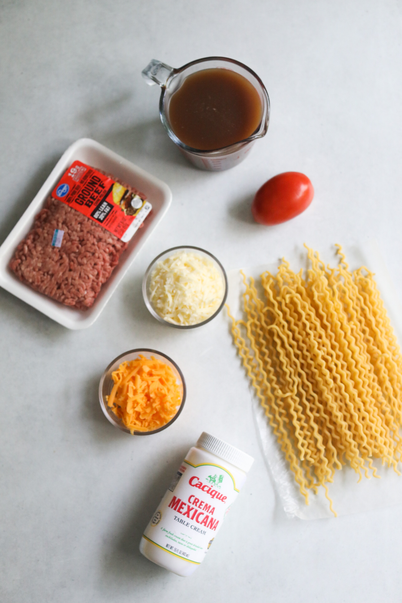 One pot taco spaghetti ingredients flat lay for additional ingredients. Packaged ground beef, beef stock in a measuring glass, roma tomato, two bowls with cheddar and Monterey jack cheese, Mexican sour cream and noodles.