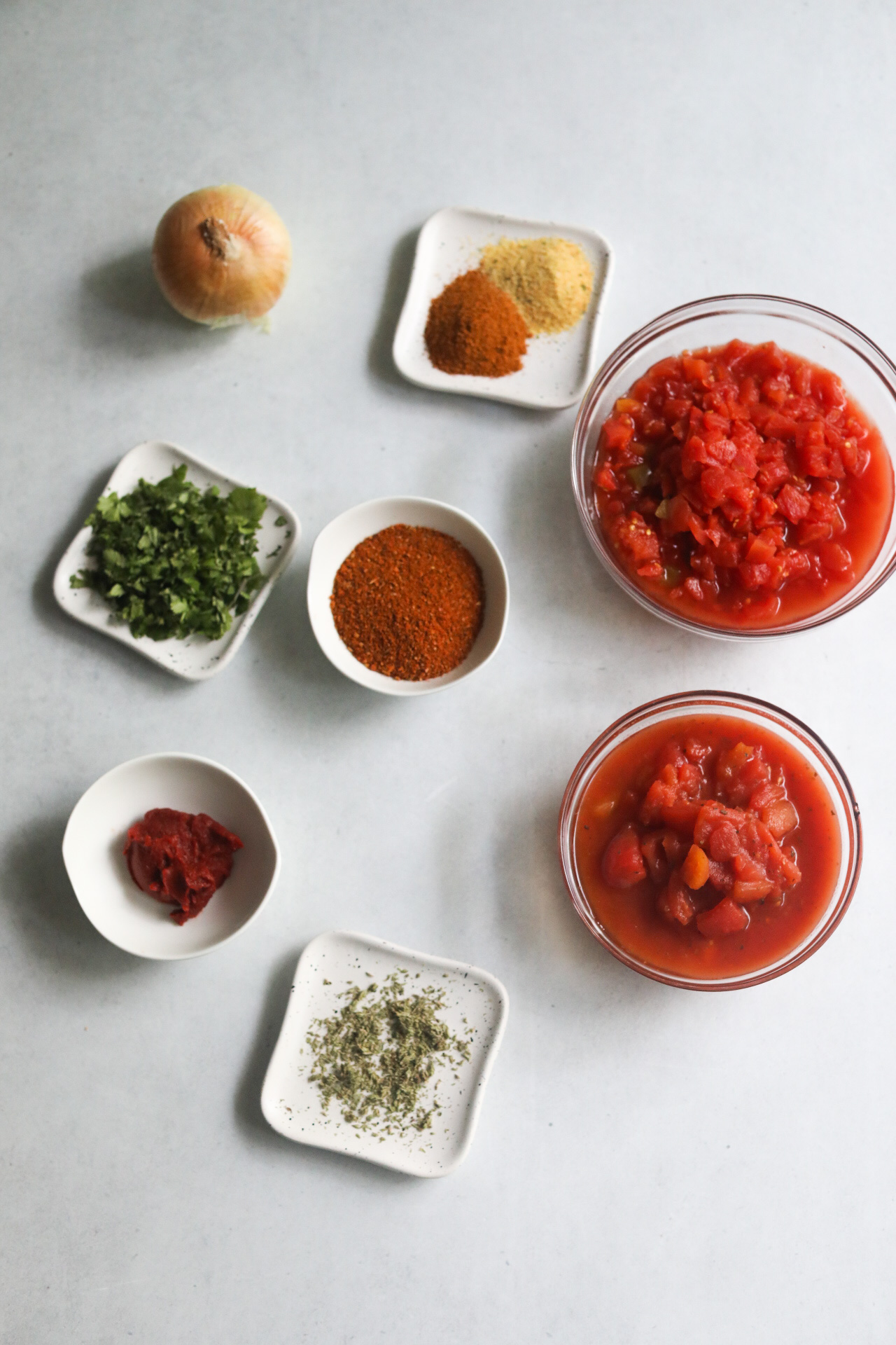 One pot taco spaghetti ingredients in a flat lay. Bowls filled with canned and diced tomatoes, seasonings, tomato paste, and herbs. A small yellow onion is also added.