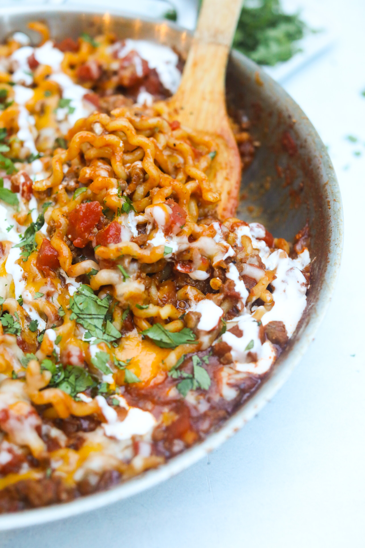 One pot taco spaghetti served in a metal cooking pan. A wooden flat spoon is scooping the pasta to show a closer view of the recipe. The pasta dish is garnished with sour cream drizzle, cilantro and diced tomatoes.