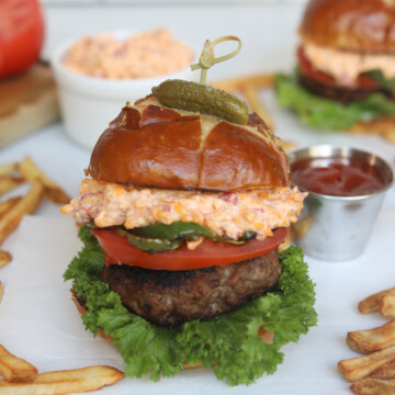 Image of pimento cheese burgers. One is in focus stacked with fries added around the burger for styling purposes. A small white ramekin filled pimento cheese and an aluminum small sauce cup filled with ketchup.