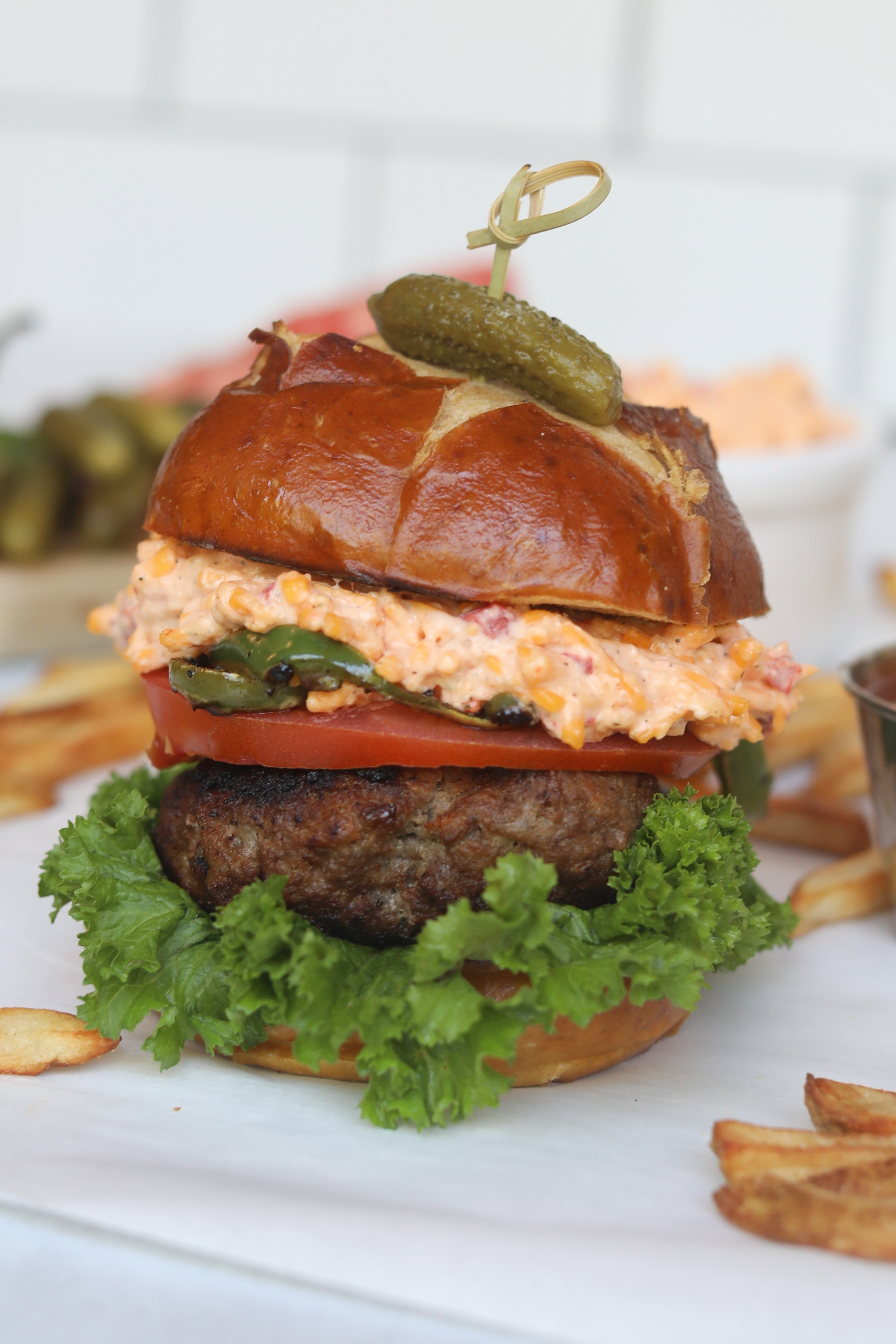 Pimento cheese burger stacked with pickle on top held by a bamboo toothpick. Fries are added around the burger for styling purposes.
