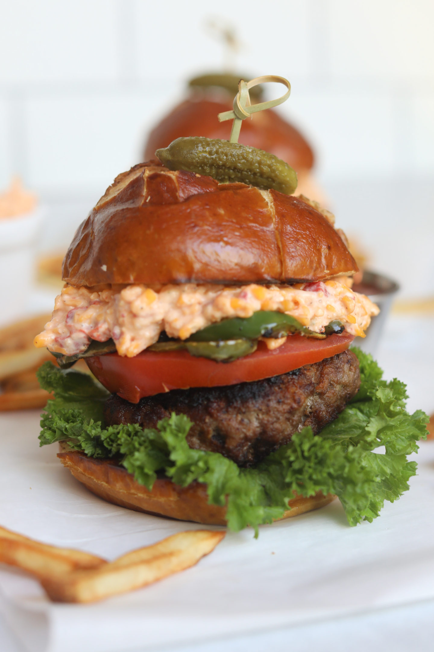 Final shot of recipe of pimento cheese burger stacked with a pickle on top held by a bamboo toothpick. Fries added around the burger for styling purposes.