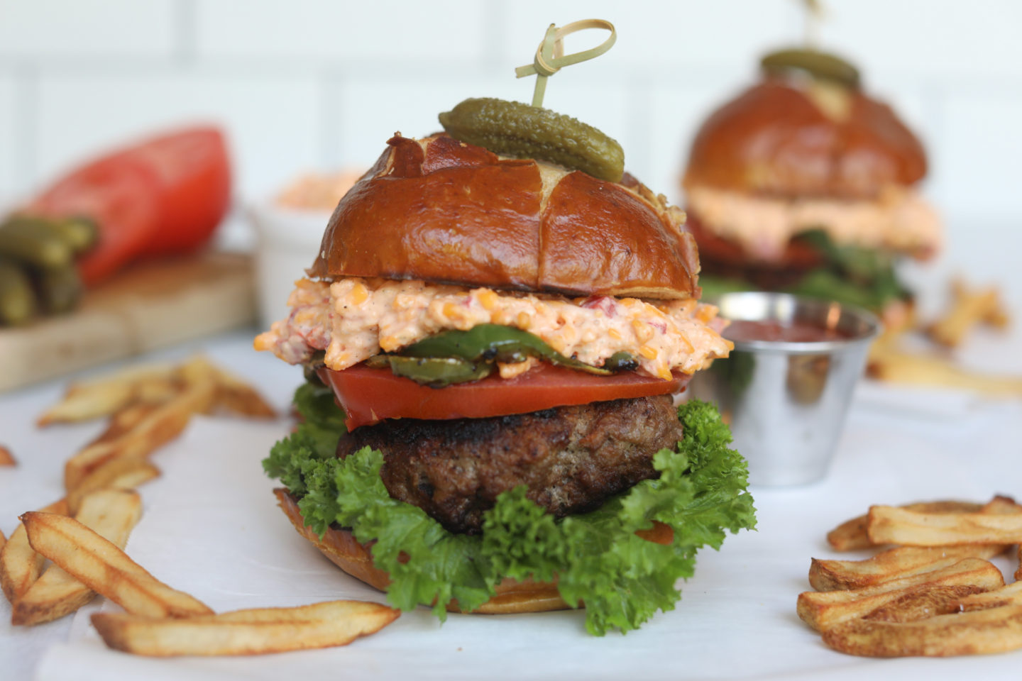 Pimento cheese burger stacked with fresh green lettuce, burger patty, sliced tomato, serrano peppers, pimento cheese topped with bun and small pickle. Fries added for styling purposes around the burger.