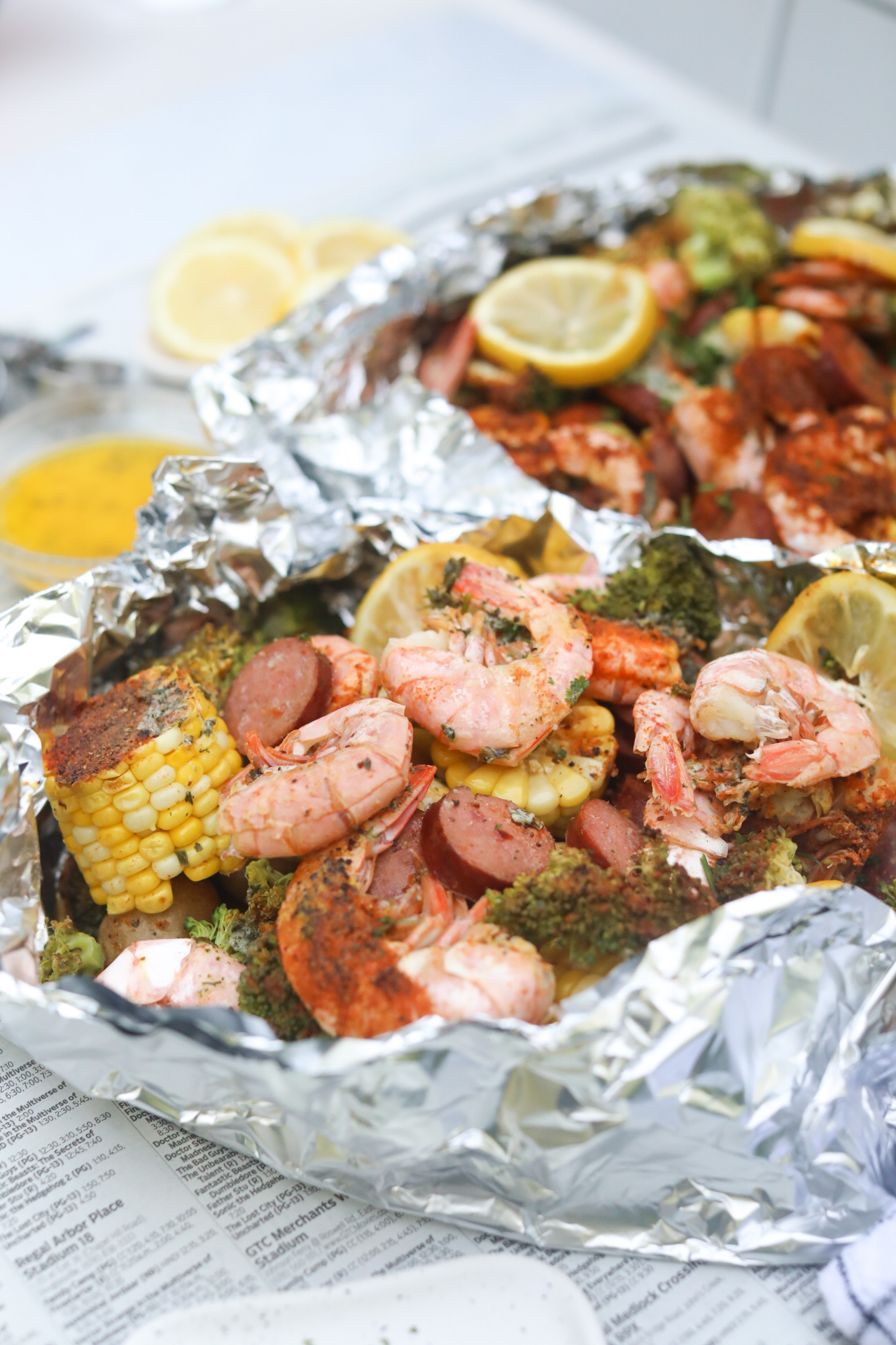 Shrimp boil in foil packed baked and top aluminum foil removed to show completed recipe. Foil packet is underneath newspaper and styled with a black and white kitchen towel. Angle is slightly from the top, revealing baked ingredients drenched in butter and seasonings.