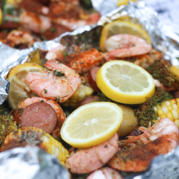 Shrimp boil in foil packed baked and top aluminum foil removed to show completed recipe. Foil packet is underneath newspaper and styled with a black and white kitchen towel. Angle is slightly from the side revealing baked ingredients drenched in butter and seasonings.