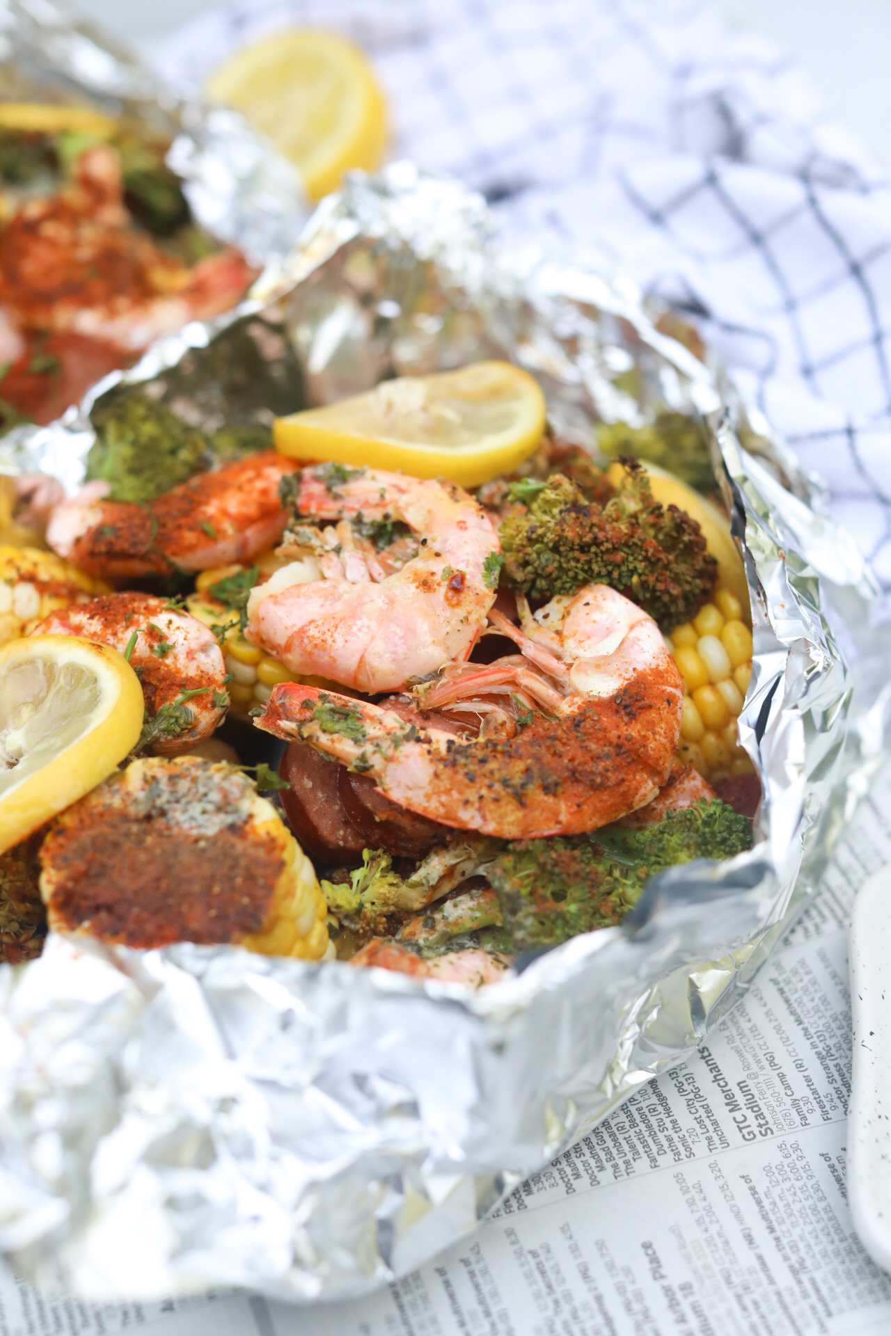 Shrimp boil in foil packed baked and top aluminum foil removed to show completed recipe. Foil packet is underneath newspaper and styled with a black and white kitchen towel.