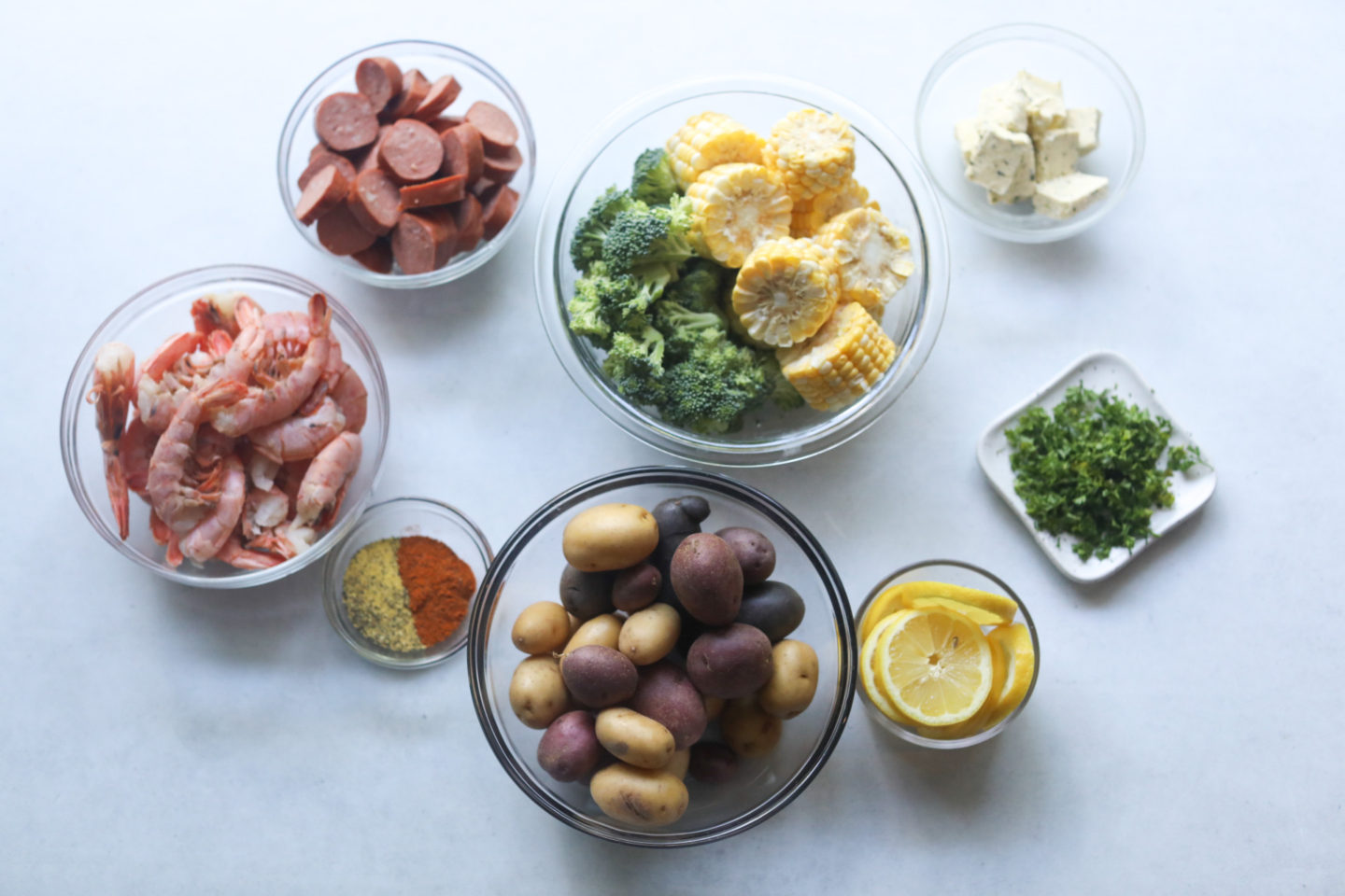 Shrimp foil packets in oven ingredients. In a flat lay are clear bowls filled garlic butter, sausage, shrimp, Cajun and lemon and pepper seasoning, small assorted colored potatoes, sliced lemons, and a small white plate with chopped parsley.