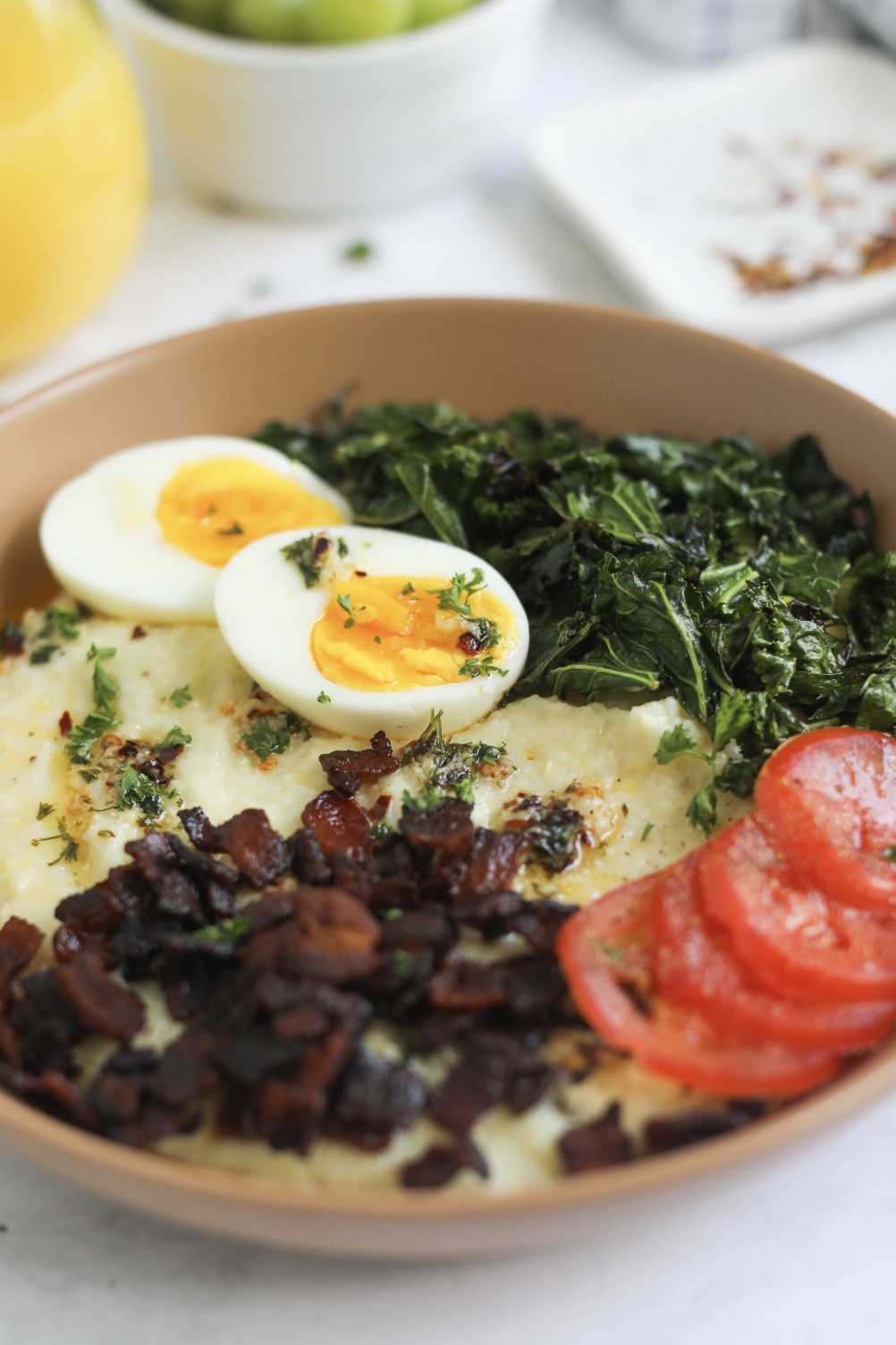 Soft boiled eggs and sauteed kale in focus of Southern Grits and Greens Bowl. Out of focus behind bowl is a glass of orange juice in a wine glass, small plate of crush red pepper flakes and a white bowl of grapes.