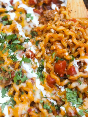 Taco spaghetti in a metal pan topped with sour cream, cilantro being scooped with a brown wooden spoon.